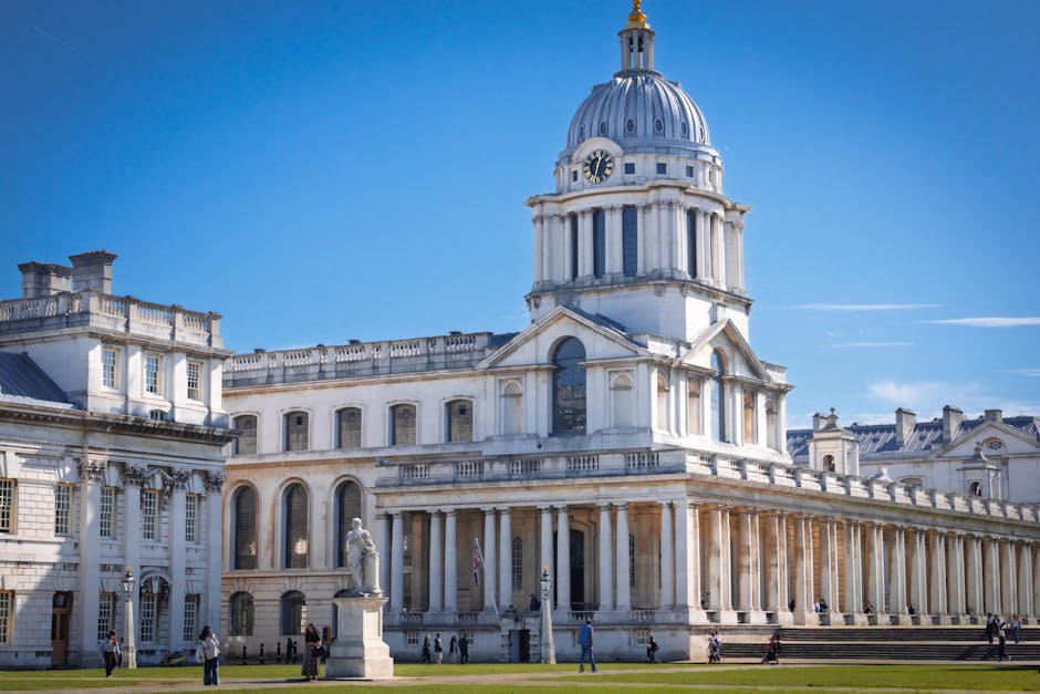 The image shows a large, historic neoclassical building with a prominent domed clock tower at the top, set against a clear blue sky. In the foreground, there is a paved area with several people walking and standing nearby, some near statues that are positioned along the building's facade. The building features tall columns supporting a pediment, with multiple arched windows on the lower level and rectangular windows on the upper levels. To the side, a portion of the building extends with more architectural details consistent with classical style. The scene appears to be captured during daylight with bright natural lighting, emphasizing the white stone construction. This setting could be associated with a well-known historic landmark or government building in the UK, and the image context aligns with themes of relocation or home transformation, subtly connecting to furniture or object transport considerations in house removals, as would be relevant to Greenwich Removals' services.