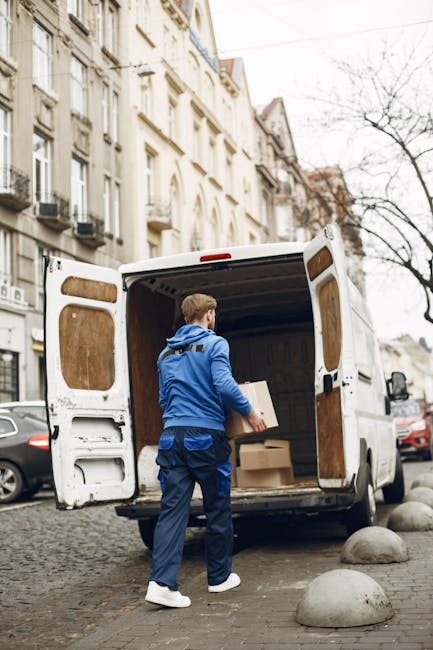 A man wearing a blue jacket, dark blue trousers, and white sneakers is seen loading cardboard boxes into the back of a white commercial van with open rear doors, parked on a cobblestone street. The van's interior is lined with wooden panels, and the boxes vary in size, with some wrapped in packing paper or plastic film. The street is lined with multi-story residential buildings featuring classic architecture, large windows, and ornate details, with other vehicles parked nearby. The scene is overcast, and leafless trees are visible in the background, indicating a cold season. Greenwich Removals is operating in this environment, supporting home relocation and furniture transport services, with the loading process taking place outside a residential property as part of a house removal or packing and moving operation.