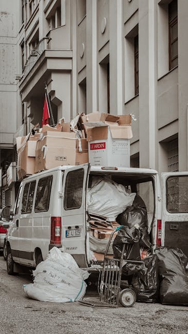 A white van parked on a street in front of a multi-storey building with a beige facade and multiple windows. The van’s rear doors are open, revealing various packed cardboard boxes, some labeled and sealed with packing tape, along with large black plastic trash bags and wrapped furniture or household items. A hand trolley and a few other packing materials are positioned outside the van, indicating a loading or unloading process typical of home relocation or furniture transport activities. The scene captures the logistics involved in a house removals service, with Greenwich Removals providing a solution for bulky item removals, as part of their professional moving and packing services in the area.