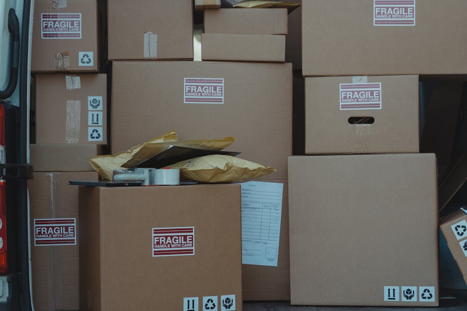 A man wearing a blue jacket, dark blue trousers, and white sneakers is seen loading cardboard boxes into the back of a white commercial van with open rear doors, parked on a cobblestone street. The van's interior is lined with wooden panels, and the boxes vary in size, with some wrapped in packing paper or plastic film. The street is lined with multi-story residential buildings featuring classic architecture, large windows, and ornate details, with other vehicles parked nearby. The scene is overcast, and leafless trees are visible in the background, indicating a cold season. Greenwich Removals is operating in this environment, supporting home relocation and furniture transport services, with the loading process taking place outside a residential property as part of a house removal or packing and moving operation.