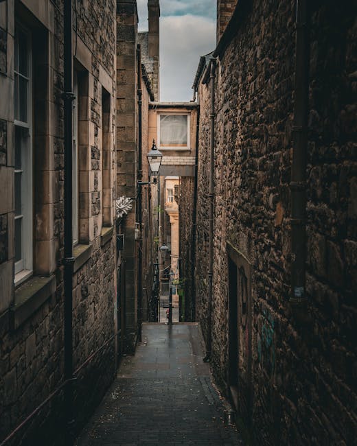 A narrow, cobbled alleyway between two old stone buildings in Greenwich, with a traditional black metal street lamp mounted on the left wall and a small window above it. Down the alley, a moving trolley loaded with cardboard boxes and wrapped furniture is visible, partially obscured by the confined space. The environment appears to be part of a home relocation process, with packing materials and belongings prepared for transport. A van or moving vehicle may be parked at the end of the alley, although it is not visible in this image. The scene is lit by natural daylight, highlighting the textured stone walls and the confined urban setting, reflecting challenges faced during furniture transport through narrow streets with Greenwich Removals providing expert moving services for such situations.