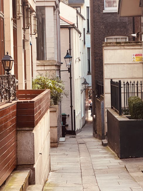 A narrow residential alleyway in Greenwich showing a paved walkway lined with buildings on both sides. On the left, there is a modern wooden planter box with plants, a decorative fence, and a vintage-style lantern mounted on a wall. On the right, a black metal fence surrounds a small garden area with shrubbery, and a building with a brick façade and a yellow warning sign. Further down the alley, a person is partially visible carrying a large cardboard box, with a moving truck or van visible in the background, indicating a home relocation or furniture transport process. The scene is lit by natural daylight, with multiple street lamps mounted along the buildings, contributing to the atmosphere of a quiet, urban neighbourhood, typical for house removals in tight street environments, as discussed in Greenwich Removals’ insider tips for narrow streets.