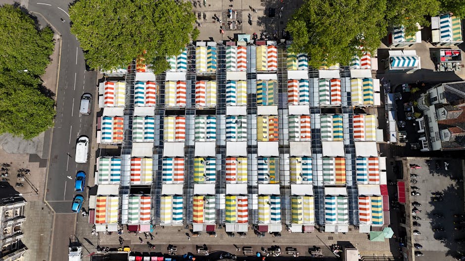 An aerial view of numerous market stalls set up in a city square, each covered with brightly coloured striped canopies in shades of red, yellow, blue, green, and white. The stalls are arranged in a grid formation, with narrow pathways between them, and some vendors and visitors are visible walking around the area. Surrounding the market are trees with lush green foliage, adjacent buildings, and a street with parked cars on the left side of the image. In the background, the tops of additional structures and more pedestrian activity can be seen. This scene captures a busy outdoor market environment, typical of a home relocation or moving process that requires careful packing and transport of furniture and goods, similar to the services offered by Greenwich Removals. The lighting is natural, indicating daytime with clear visibility of all objects and details.