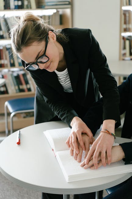 A woman wearing glasses and a black blazer is leaning over a round white table, helping to lift or move a cardboard box wrapped in clear plastic. She is in a room with bookshelves filled with books in the background, and natural light illuminates the space. On the table, there is a red pen and some documents or packing materials. The scene depicts a team member of Greenwich Removals during a home relocation process, possibly preparing items for packing or transportation as part of a furniture transport or moving service, emphasizing careful handling and packing in the context of a house removal within the Greenwich area.