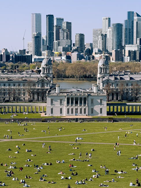 A large white building with classical architectural features, including columns and twin towers with domed roofs, situated on a green lawn with numerous people sitting and lying on the grass. The scene is set in a park with pathways and trees, and behind the park, there is a river and a city skyline featuring tall modern skyscrapers made of glass and steel. The photograph captures the outdoor setting during daytime with clear weather. Greenwich Removals' home relocation and furniture transport services may involve similar outdoor loading and unloading activities, with crews managing packing and moving logistics in urban and park environments.