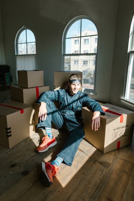 A young man sitting on a wooden floor inside a room with white walls and large arched windows, surrounded by multiple packed cardboard boxes sealed with red and black packing tape. The boxes are stacked and arranged around him, indicating a house move or packing process. The sunlight streams through the windows, illuminating the space and highlighting the boxes and the man's casual, relaxed posture. This scene reflects the preparation and logistics involved in home relocation, with Greenwich Removals providing professional furniture transport and packing services for a seamless move in Greenwich.