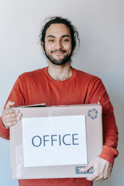 In the image, a man with dark, curly hair and a beard is standing indoors against a plain light gray wall. He is wearing a red long-sleeved shirt and is smiling gently while holding a cardboard box labeled 'OFFICE' with both hands. The box appears to be part of a professional office relocation process, reflecting aspects of packing, furniture transport, and moving logistics. The scene suggests a home or office environment where professional removals services, such as those offered by Greenwich Removals, are involved. The lighting is even and natural, highlighting the details of the box and the man's expression, emphasizing the careful handling of office items during a house or business move.