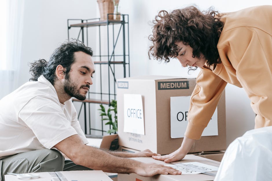In the image, a man with dark, curly hair and a beard is standing indoors against a plain light gray wall. He is wearing a red long-sleeved shirt and is smiling gently while holding a cardboard box labeled 'OFFICE' with both hands. The box appears to be part of a professional office relocation process, reflecting aspects of packing, furniture transport, and moving logistics. The scene suggests a home or office environment where professional removals services, such as those offered by Greenwich Removals, are involved. The lighting is even and natural, highlighting the details of the box and the man's expression, emphasizing the careful handling of office items during a house or business move.
