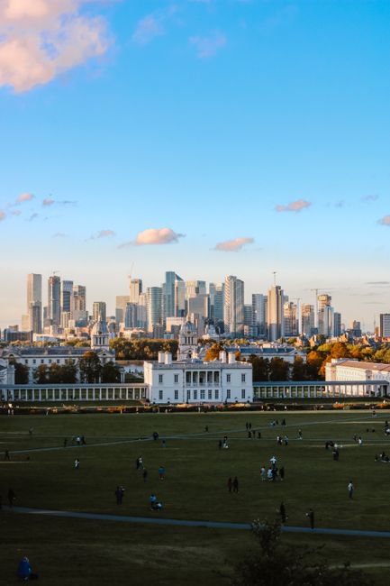 A wide shot of a city skyline during daytime with tall modern office buildings and skyscrapers in the background under a blue sky with scattered clouds. In the foreground, there is a large open park with a well-maintained grassy area, pathways, and numerous people walking, sitting, and enjoying leisure activities. The park features a white colonial-style building facing the city, situated on the edge of the green space. This scene captures the contrast between urban development and recreational outdoor space, suitable for illustrating home relocation or moving services associated with furniture transport and packing processes, as offered by Greenwich Removals. The location appears to be in a major metropolitan area, highlighting logistics involved in city house removals and property moves.
