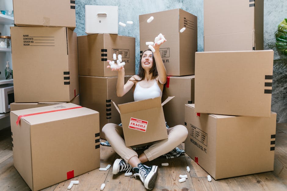A young woman with long dark hair, dressed in a white tank top and beige pants, is sitting cross-legged on a wooden floor inside a room during a home relocation process. She is smiling and looking upward while tossing white packing foam peanuts from an open cardboard box onto the floor. Surrounding her are several large cardboard moving boxes, some sealed with red tape and others open, revealing additional packing materials and smaller boxes inside. The boxes are stacked against a textured wall, with a small potted green plant visible in the corner. To her left, a portion of a white microwave and kitchen countertop can be seen, indicating the room may be part of a residential kitchen or living area. Behind her, a door or window frame is partially visible, and the lighting appears natural, highlighting the scene of packing and furniture transport preparation in a typical house removal from Greenwich Removals. The overall setting suggests a systematic packing and loading process involved in furniture transport or house clearance, with essential equipment like packing materials and carefully stacked boxes visible throughout.