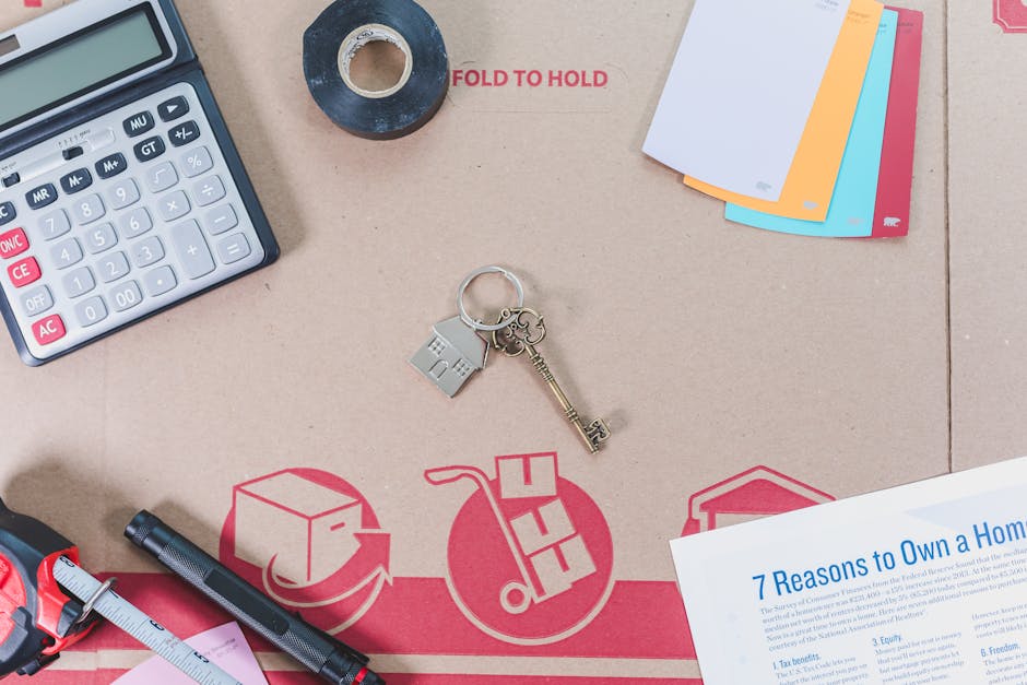 Top-down view of a light brown cardboard box used for home relocation, with various packing and moving essentials on its surface. Visible items include a calculator with large white and red buttons, a roll of black tape, a set of multicoloured plastic folders, a keyring with a small house-shaped keychain and a metallic key, a black marker pen, a pink measuring tape, and a printed sheet of paper titled '7 Reasons to Own a Home.' The cardboard box features printed graphics of a moving cart and a package, indicating its use for removals or furniture transport. The scene captures the packing and organisation process involved in house removals, highlighting tools and materials typically used by Greenwich Removals and other professional moving services.
