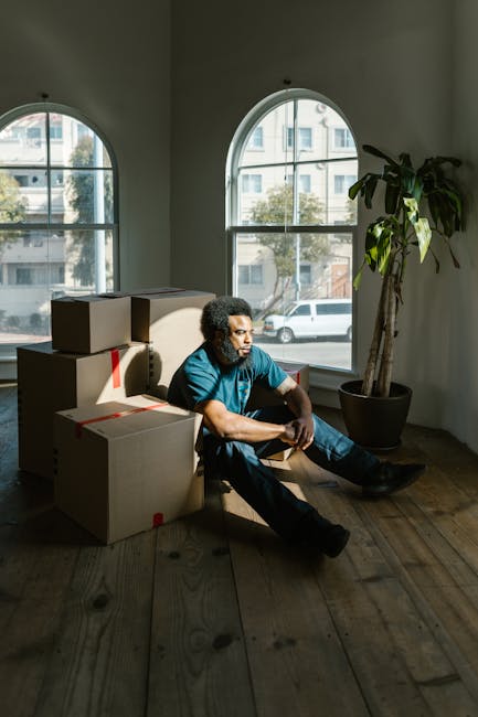 A man with curly dark hair and a beard, dressed in a blue T-shirt and dark trousers, sitting on a wooden floor inside a room with two large arched windows that let in natural light and reveal a street scene with parked cars and buildings outside. He is surrounded by several cardboard moving boxes, some stacked, with red tape seals and partially open flaps, indicating packing for a house relocation or moving process. The boxes are placed near a modern potted plant with large green leaves, and the interior space features white walls and wooden flooring, suggesting the setting is part of a home or apartment ready for a move. The scene depicts the preparation stage of furniture transport and packing and moving tasks, which Greenwich Removals may assist with, aligning with the services described on the page about flat removals costs and quotes.