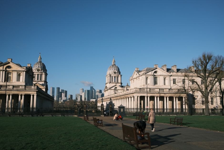 A wide-angle view of a historic architectural complex featuring classical-style buildings with domed cupolas, situated in an urban park setting during daytime. The foreground displays a paved pathway with wooden benches on either side, where a person in a grey jacket is walking along, and a dog is being held on a leash. A person is seen bending over on a park bench, possibly arranging or inspecting packing materials, such as cardboard boxes and plastic wrap, positioned nearby. The scene includes outdoor loading equipment like a trolley or cart used in furniture transport or home relocation activities. Tall trees with sparse branches stand to the right, and the background features modern high-rise buildings, indicating a city center. The lighting suggests clear weather with bright sunlight illuminating the scene, emphasizing the process of packing or loading furniture and boxes for a house move, as part of a professional removals service by Greenwich Removals. The setting aligns with the topic of house removals and parking regulations for relocations in Greenwich, London.