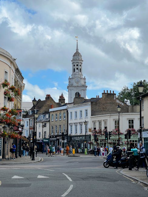 A busy street scene in Greenwich, featuring a row of multi-story buildings with various facades, some adorned with flower baskets and outdoor seating areas. Several pedestrians are walking along the sidewalks, and a few people are gathered near shop entrances. On the right side, a black motorcycle is parked, and a taxis and cars are visible in motion on the road. In the background, the prominent clock tower with a spire and a weather vane stands tall against a partly cloudy sky, with patches of blue visible through the clouds. The street is well-lit, and the surroundings include street lamps and signage, reflecting a lively and well-maintained residential and commercial area within Greenwich. Greenwich Removals offers professional house removal and furniture transport services, which are relevant to efficient packing and moving processes in the area.